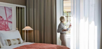 Eine Dame steht mit einem Kaffee in der Hand am Fenster ihres Hotelzimmers